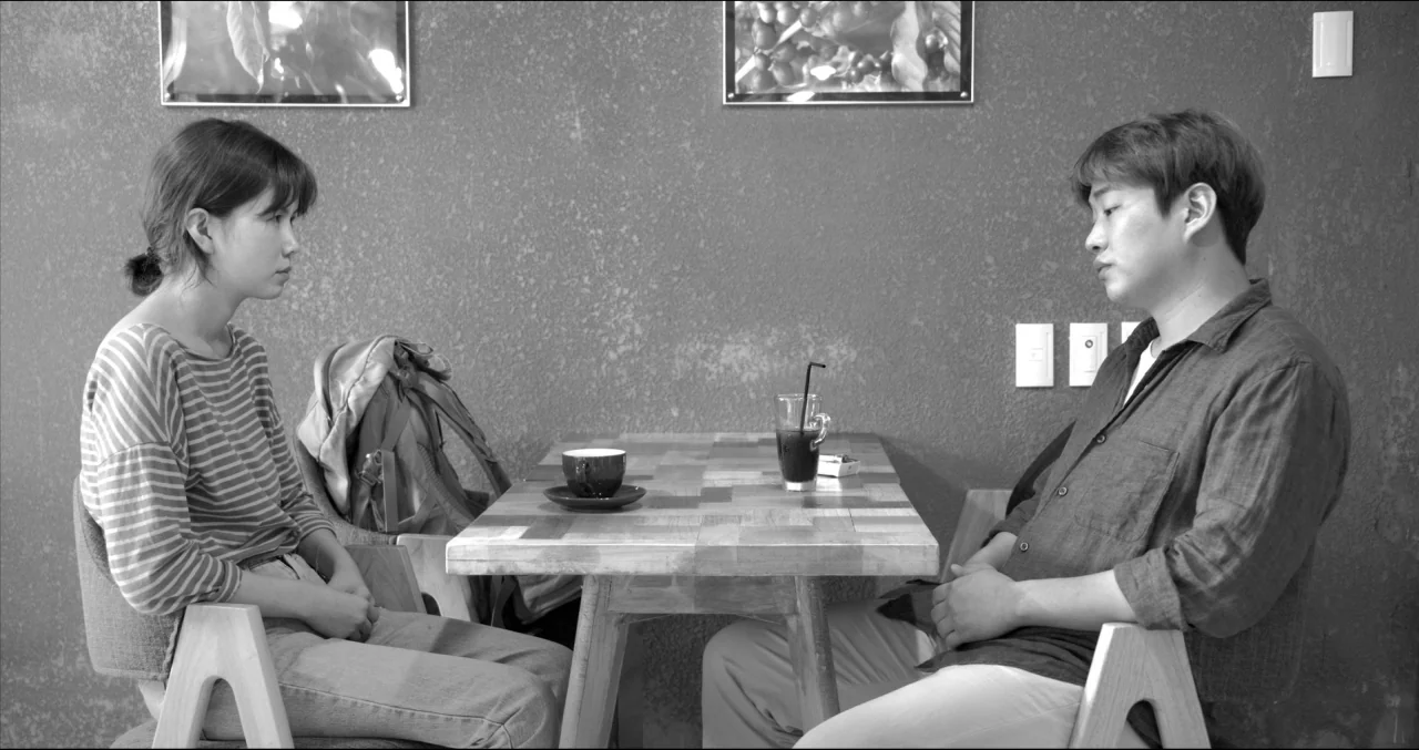 Grass (2018) movie still: medium — A man and a woman sit opposite each other at a small wooden cafe table. On the textured gray wall behind them…