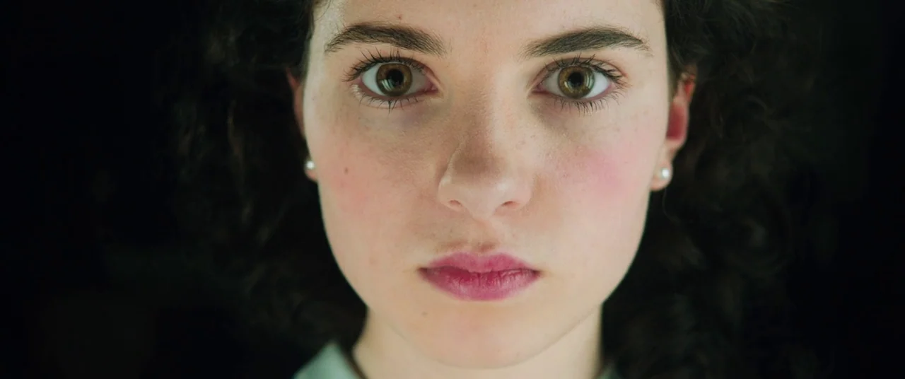 Judy (2019) movie still: closeup — A dark, featureless environment frames the subject from the shoulders up. A young woman looks directly toward the…