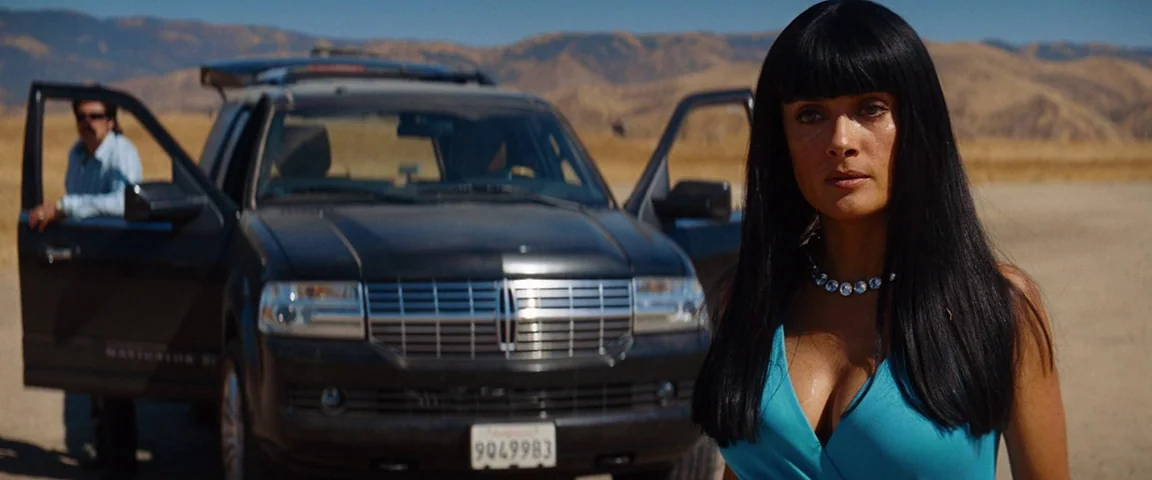 Savages (2012) movie still: medium — A dry desert landscape with rolling hills beneath a clear sky in daytime. A woman in a blue dress stands…