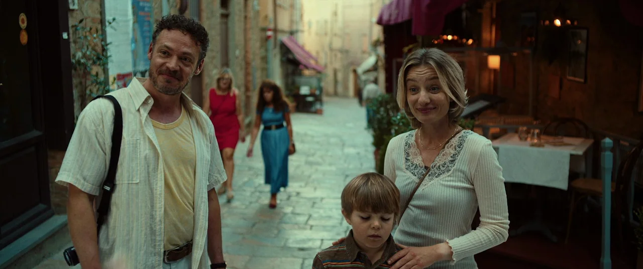 Speak No Evil (2022) movie still: medium — An old town alleyway features uneven stone pavement flanked by shop entrances and tables. A family of…