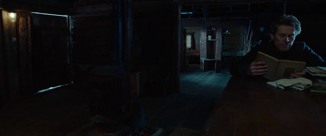 Siberia (2019) movie still: medium — A dimly lit rustic wooden cabin features log walls and a cast iron stove. An elderly man sits alone at a table…