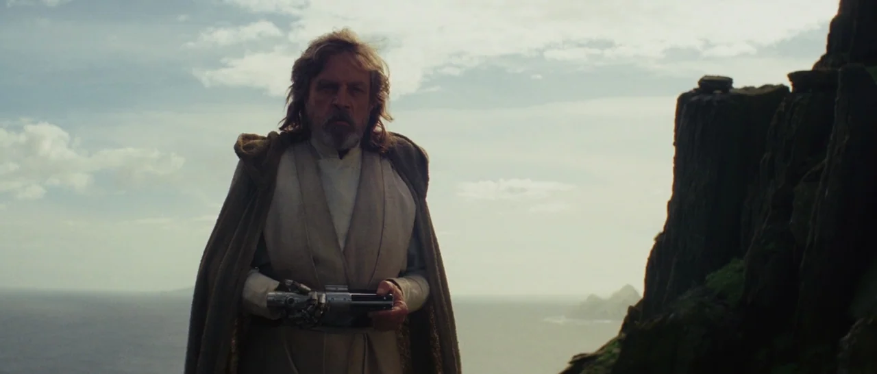 Star Wars: The Last Jedi (2017) movie still: medium — A man stands on a rocky cliff overlooking a vast sea under overcast skies. He holds a…