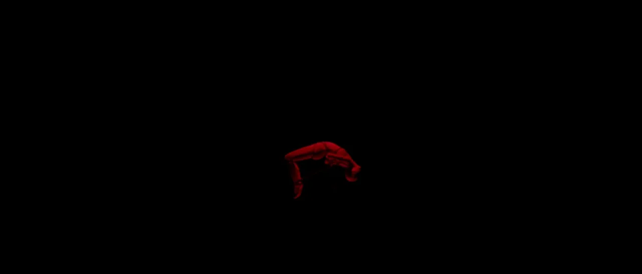 The Neon Demon (2016) movie still: wide — A figure performs a backbend in a void of total darkness. The person holds a bar while suspended in the…