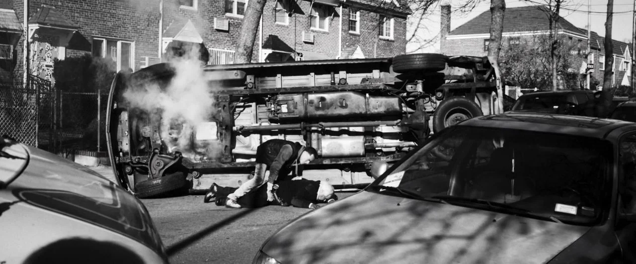 Bushwick (2017) movie still: wide — A residential city street features a large van overturned on its side with smoke. One man bends over another…