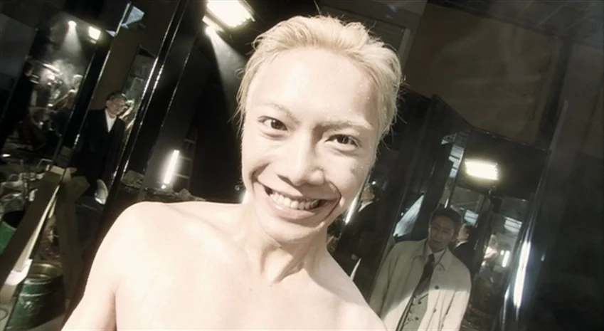 Rampo Noir (2005) movie still: closeup — A man stands in a brightly lit, sterile room with black partitions in the background. The shirtless man…