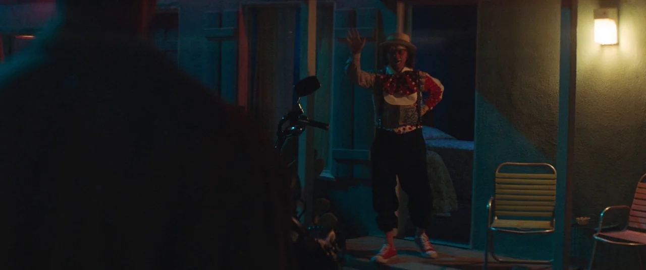 Honey Boy (2019) movie still: fullbody — A dimly lit motel walkway at night features a clown standing in a doorway. The clown waves while a…