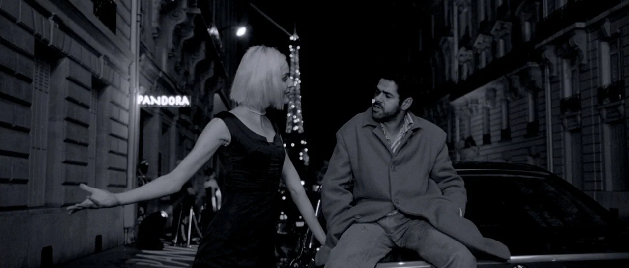 Angel-A (2005) movie still: medium — A Parisian street at night features a neon sign and the Eiffel Tower background. A blonde woman gestures to a…
