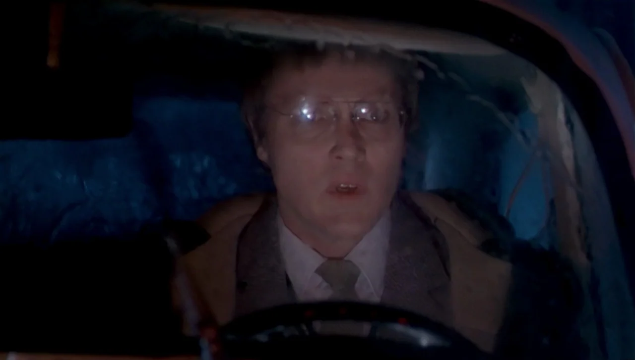 The Dead Zone (1983) movie still: closeup — A dark vehicle interior with blue night lighting reflecting off the windshield glass. A man with glasses…