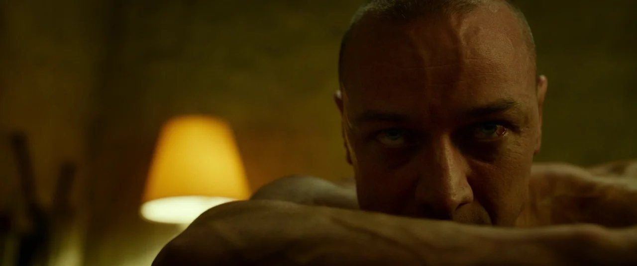 Split (2016) movie still: closeup — A dimly lit interior space features a man leaning forward towards the viewer. The background is obscured by…