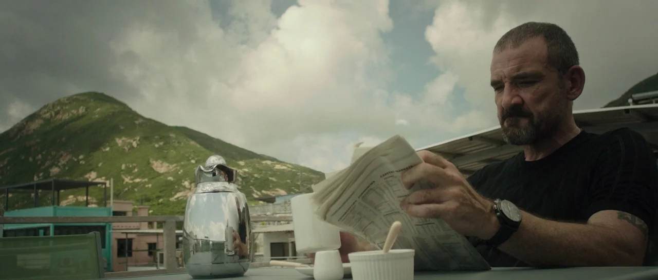 Blackhat (2015) movie still: medium — A man reads a newspaper on a terrace with rolling green hills in background. A reflective metal carafe sits on…