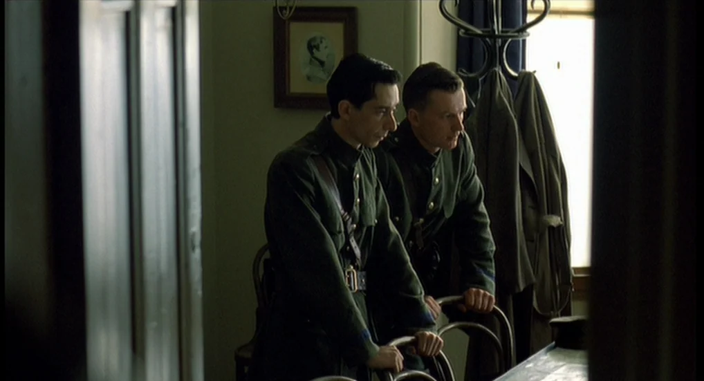 The Wind That Shakes the Barley (2006) movie still: medium — A dimly lit room with vintage furniture and a framed picture on the wall. Two men in…