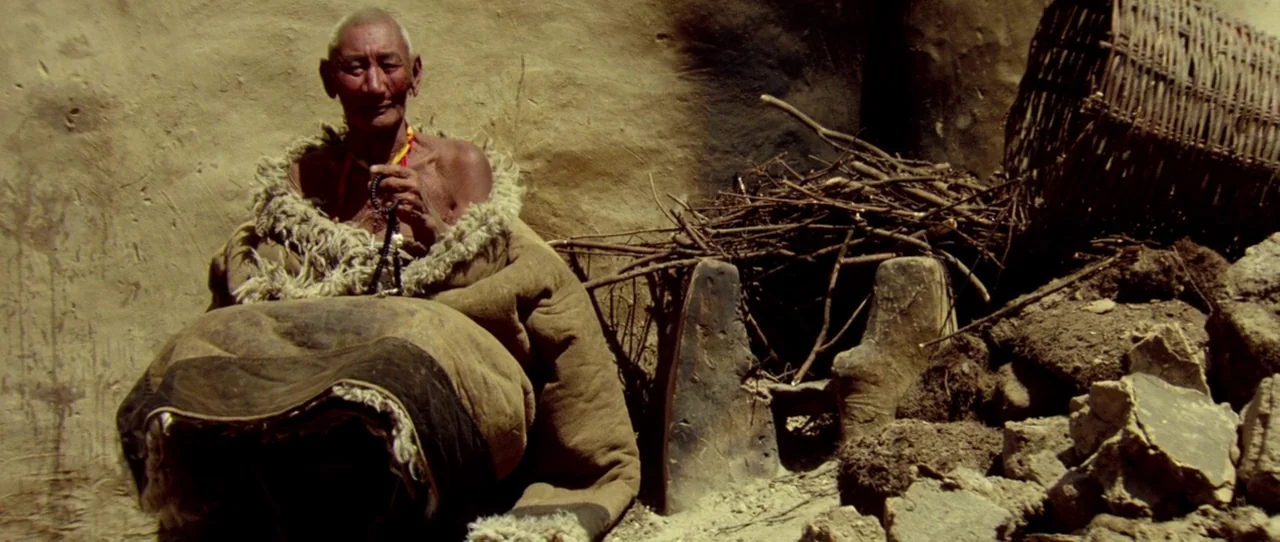 Horse Thief movie still: medium — An elderly person sits against a rough, textured mud wall in a rustic setting. They wear a heavy, worn woolen…