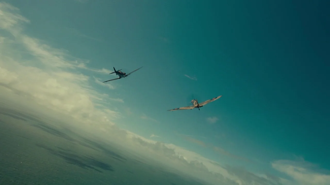 Dunkirk (2017) movie still: wide — Two vintage fighter planes fly over an expansive ocean under a partially cloudy sky. The camera perspective is…