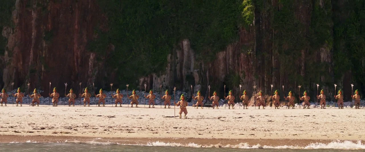 Wonka (2023) movie still: wide — A long line of identical small figures stands on a sandy beach. A rocky cliff wall rises directly behind the line…