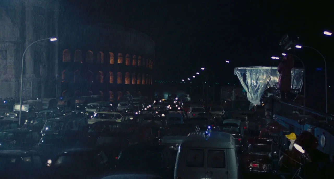Roma (1972) movie still: establishing — A dense congestion of cars fills a rainy city street at night under streetlights. The Colosseum stands…
