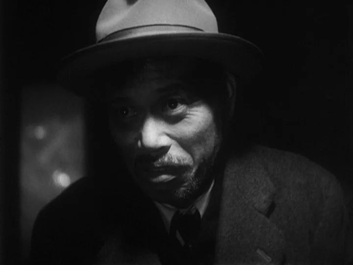 Ikiru (1952) movie still: closeup — A dark, dimly lit indoor space with low-key illumination on a man's face. The man wears a fedora and a suit…