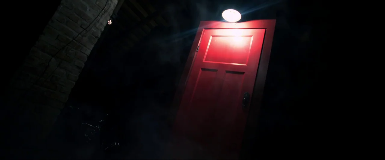 Insidious (2010) movie still: wide — A vibrant red door stands in a dark space with a brick pillar nearby. Wisps of fog drift around the door…