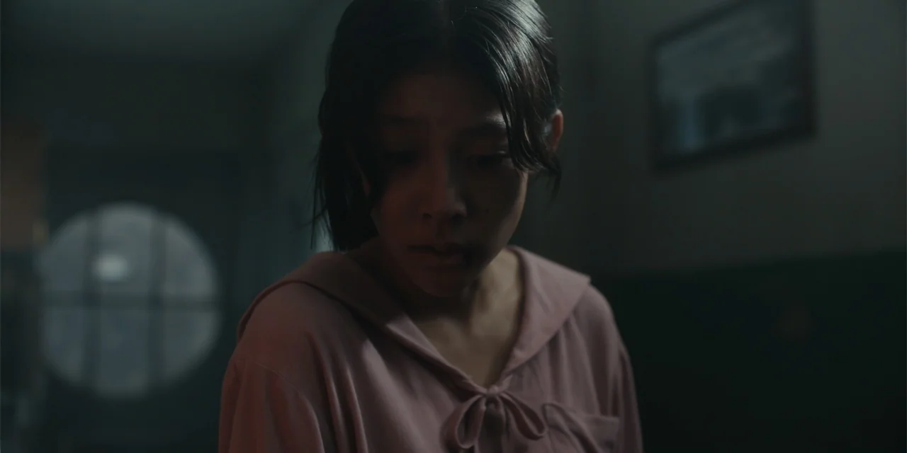 Host (2020) movie still: closeup — A dimly lit room contains a young woman standing in front of a window. She wears a pink top and looks down at an…