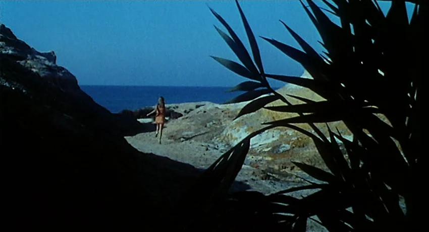 5 Dolls for an August Moon (1970) movie still: wide — A rocky coastal path overlooks a dark sea under a blue hour sky. A woman walks quickly away…