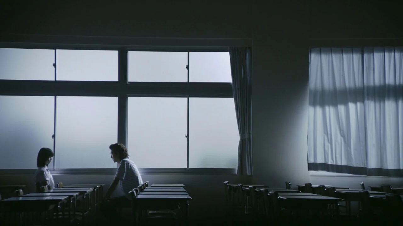 Confessions (2010) movie still: wide — A dimly lit classroom features rows of empty desks facing large, frosted windows. Two students sit at their…