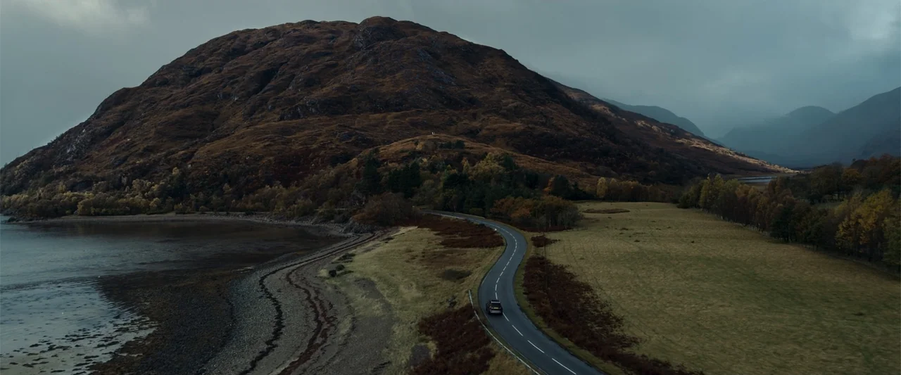 My Son (2021) movie still: establishing — A dark, winding road stretches through a coastal valley beneath large, barren mountains. A lone vehicle…