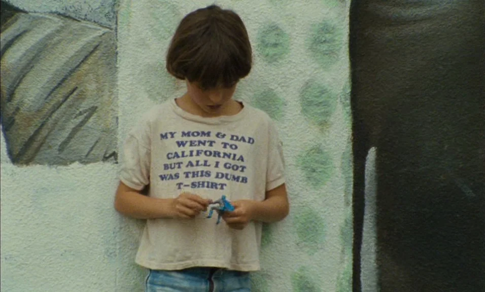 Documenteur (1981) movie still: medium — A child leans against a textured white wall featuring green and gray mural painting. The child holds a…