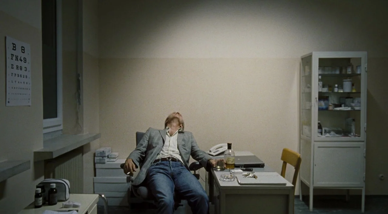 Paradise: Hope (2013) movie still: medium — A clinical examination room contains a metal desk, a glass cabinet, and eye charts. A man sits in a…