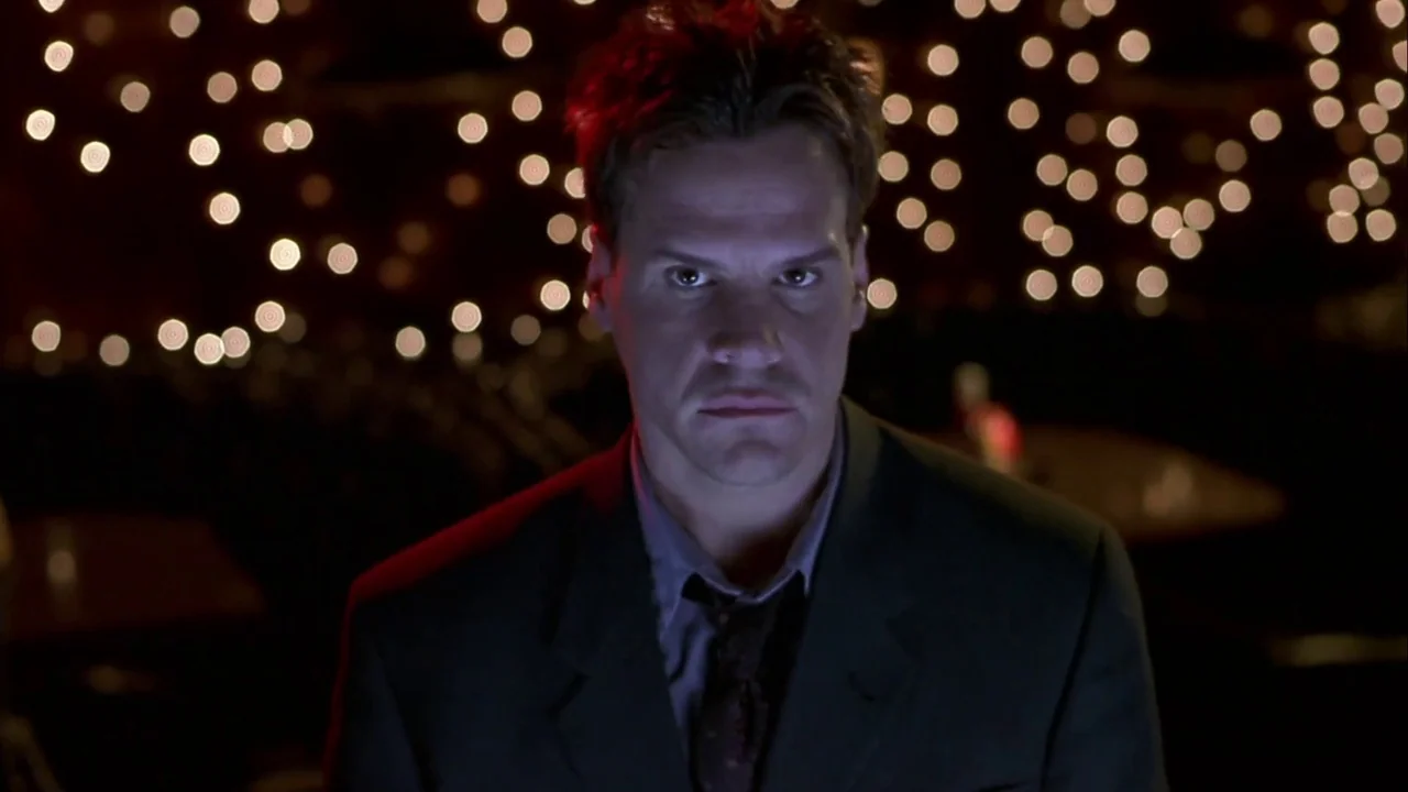 Hellraiser: Inferno (2000) movie still: closeup — A dark, dimly lit venue is filled with circular bokeh light patterns in the background. A man in a…