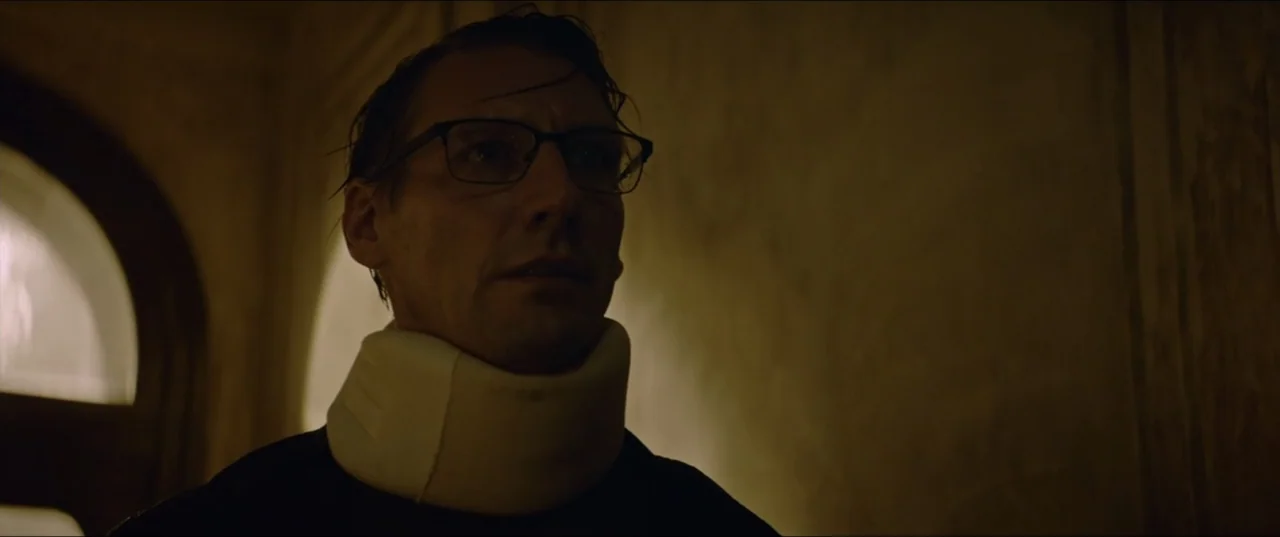 Dogs Don't Wear Pants (2019) movie still: closeup — A man wearing a neck brace stands in a dark indoor hallway. He looks toward the side while…