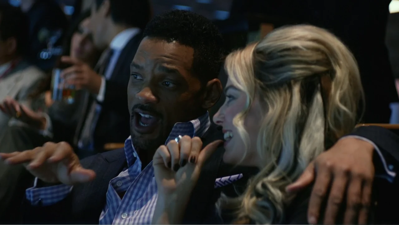 Focus (2015) movie still: medium — A crowded venue with rows of dark seats filled with spectators in the background. A man speaks animatedly to a…