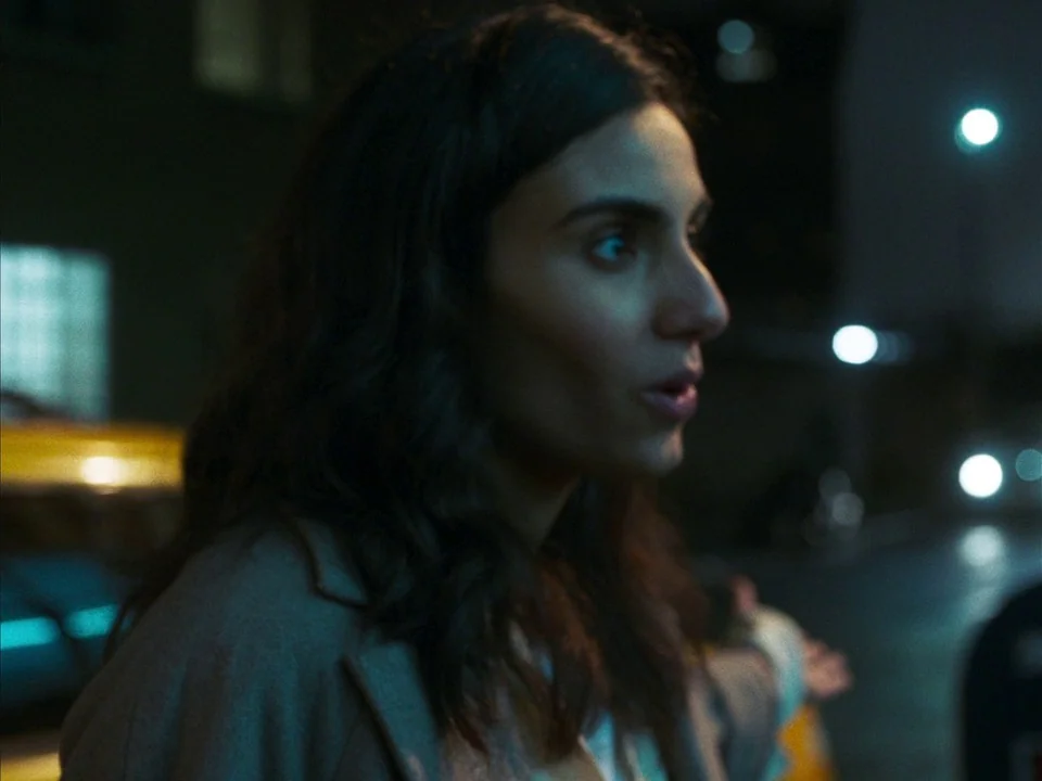 Mogul Mowgli (2020) movie still: closeup — A city street at night with blurry bokeh from lights in the background. A woman in a grey coat looks…