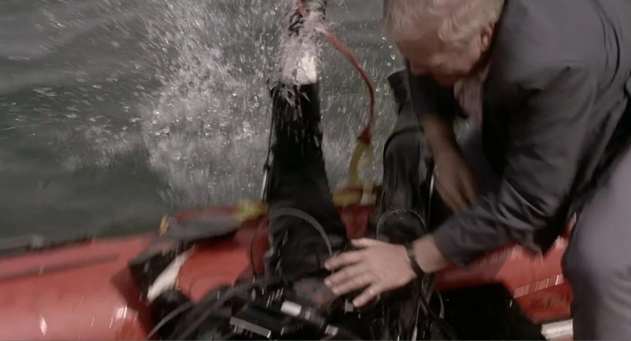 F/X2 (1991) movie still: medium — A man in a blazer leans into an inflatable boat on the water. He reaches out toward a diver wearing a wetsuit…