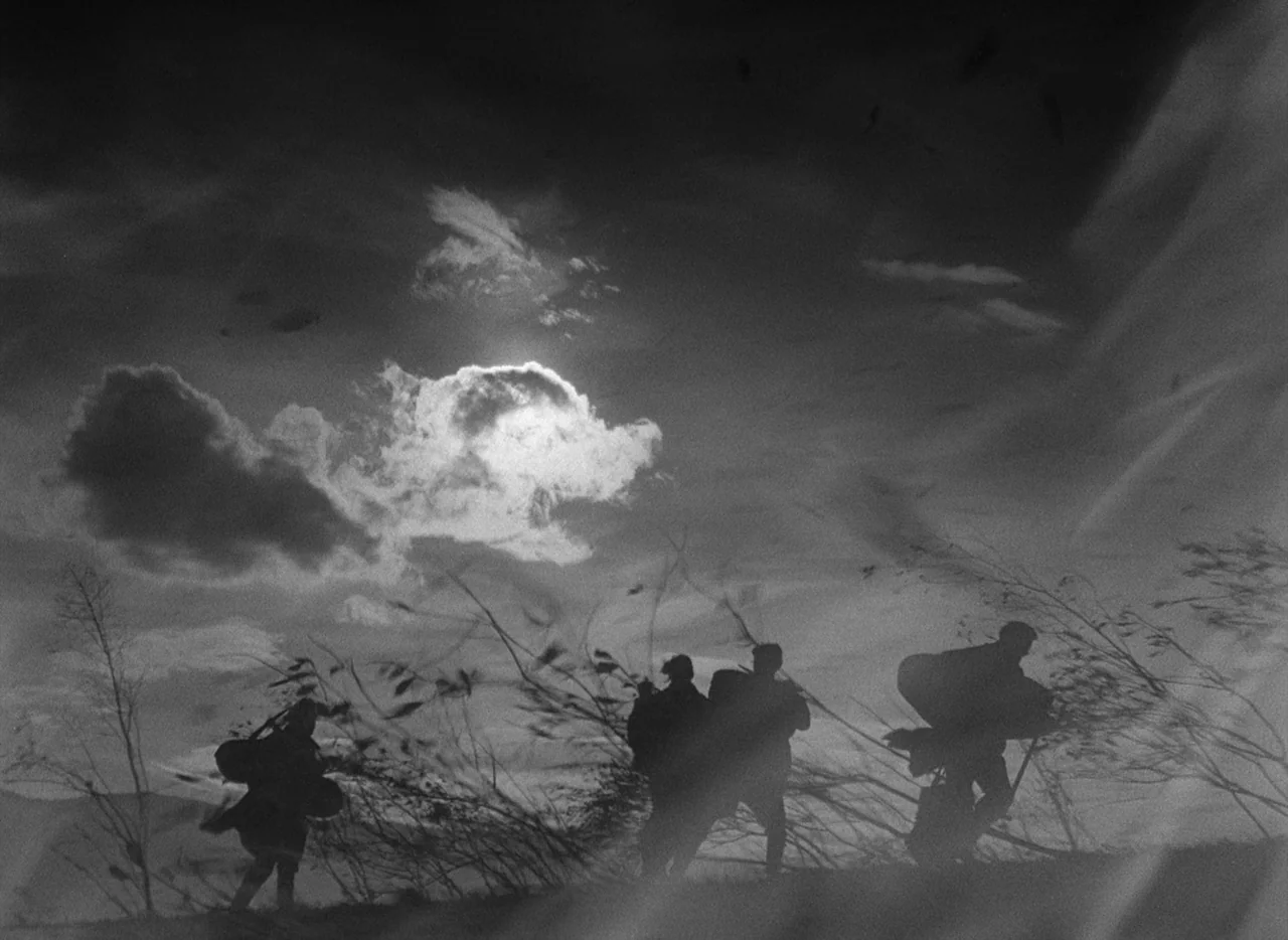 Letter Never Sent (1960) movie still: wide — A field filled with tall grass beneath a dramatic sky with sun-lit clouds. Three figures walk away from…