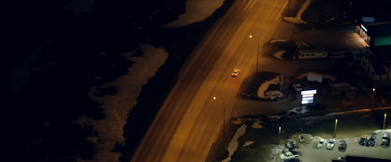 The Unknown Country (2022) movie still: establishing — A nighttime aerial view of an empty two-lane highway flanked by patches of snow. A single…