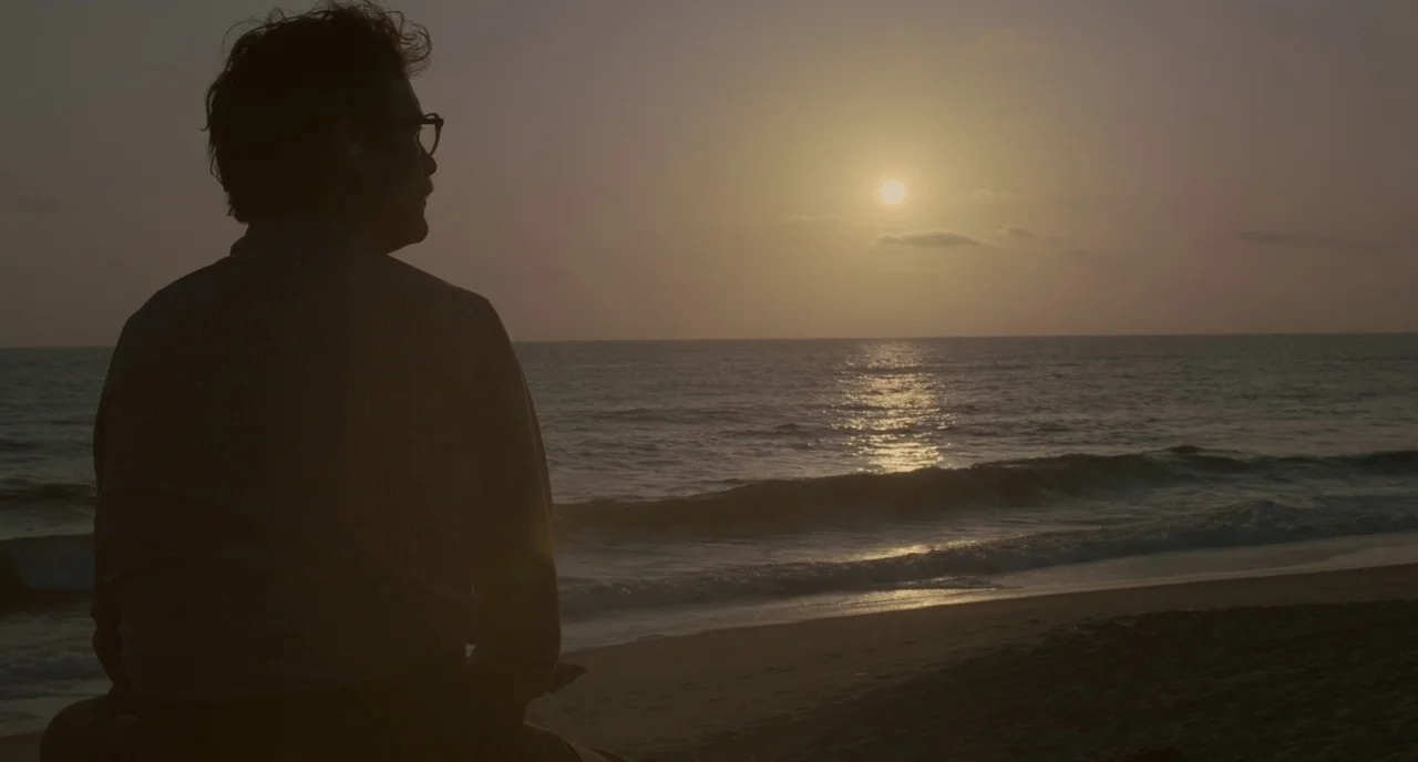 Her (2013) movie still: medium — A sandy beach shoreline at sunset features soft waves meeting the dark wet sand. A man with wire-rimmed glasses…