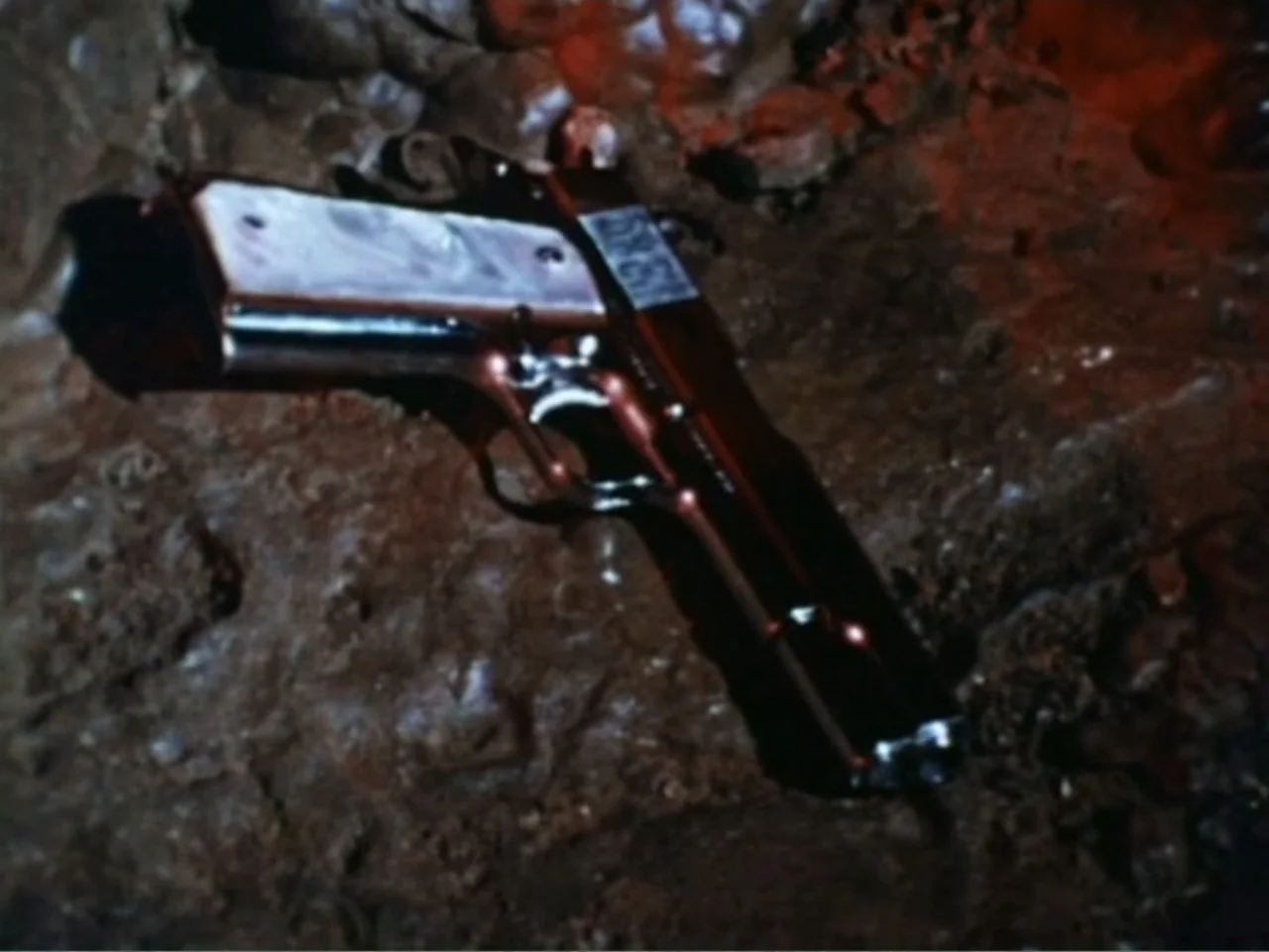 It's Alive (1974) movie still: closeup — A dark, uneven stone surface covered in wet mud and dark grit. A semi-automatic pistol with a silver grip…