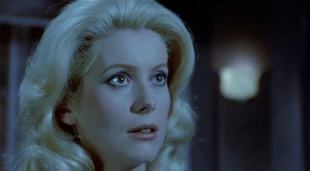 Un Flic (1972) movie still: closeup — A dark indoor setting with a blurred, illuminated wall fixture in the background. A woman with blonde hair…