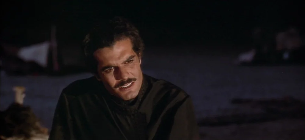 Lawrence Of Arabia movie still: medium — A dark outdoor space is lit by a low, warm light source at night. A man with a dark mustache looks toward…