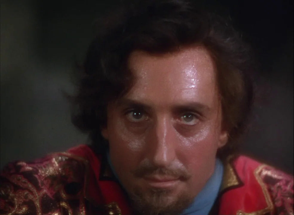 The Adventures of Robin Hood (1938) movie still: closeup — A man in ornate period clothing is centered against a dark, indistinct background. He…