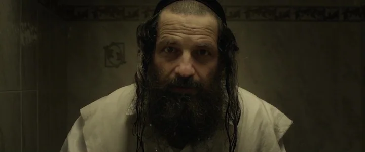 To Dust - Nightmares (2018) movie still: closeup — A tiled room with monochromatic walls forms the background. A bearded man looks forward.