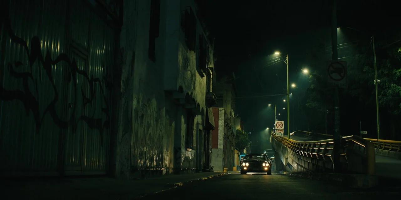 Silent Night (2023) movie still: wide — A narrow, graffiti-covered urban street stretches into the dark distance beneath overhead streetlights. A…