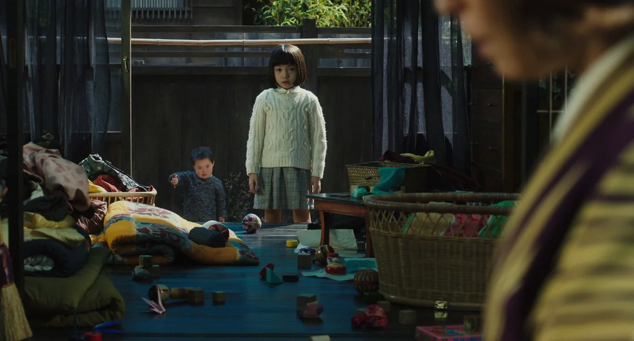 No Longer Human (2019) movie still: medium — A wooden veranda features scattered toys on the floorboards with a bamboo fence background. A young…