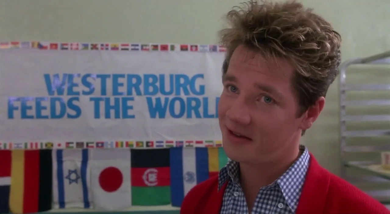 Heathers movie still: medium — A high school cafeteria interior features a white banner with blue text and flags. A young man wearing a red cardigan…