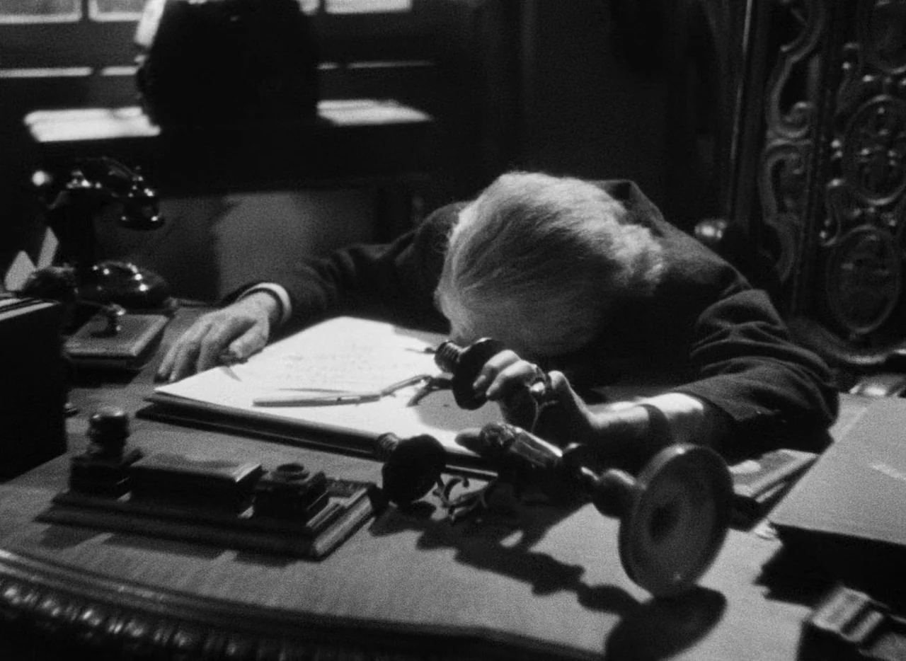 Le Corbeau (1943) movie still: medium — A dark wooden desk holds a vintage telephone and several inkwells. An elderly man in a suit is slumped…