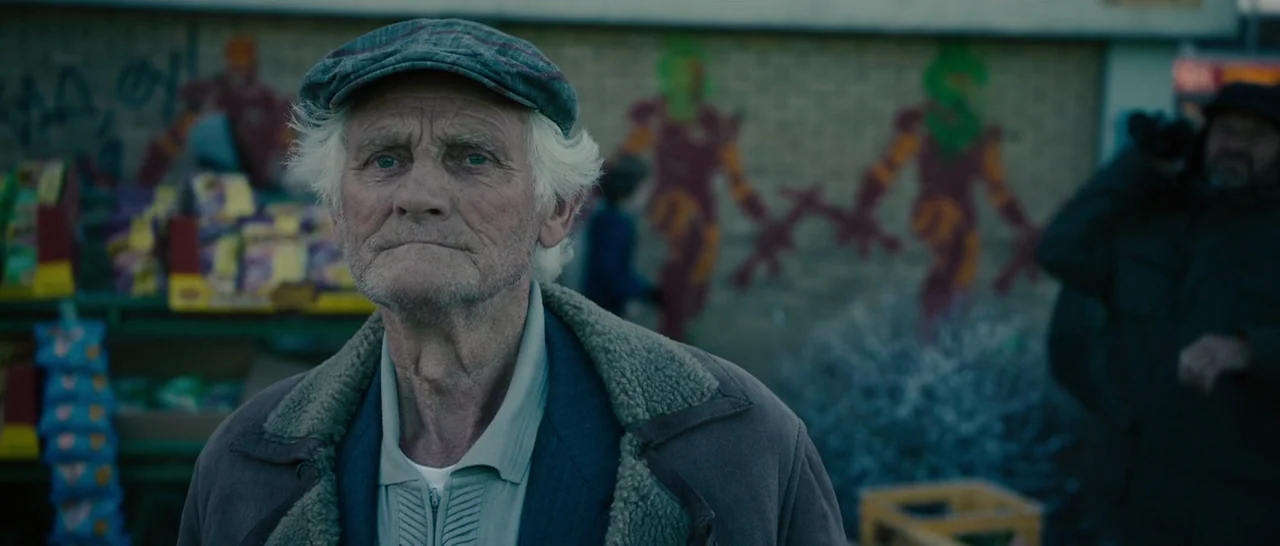 Avengers: Age of Ultron (2015) movie still: medium — An elderly man stands in front of a brick building featuring faded red graffiti. He looks…