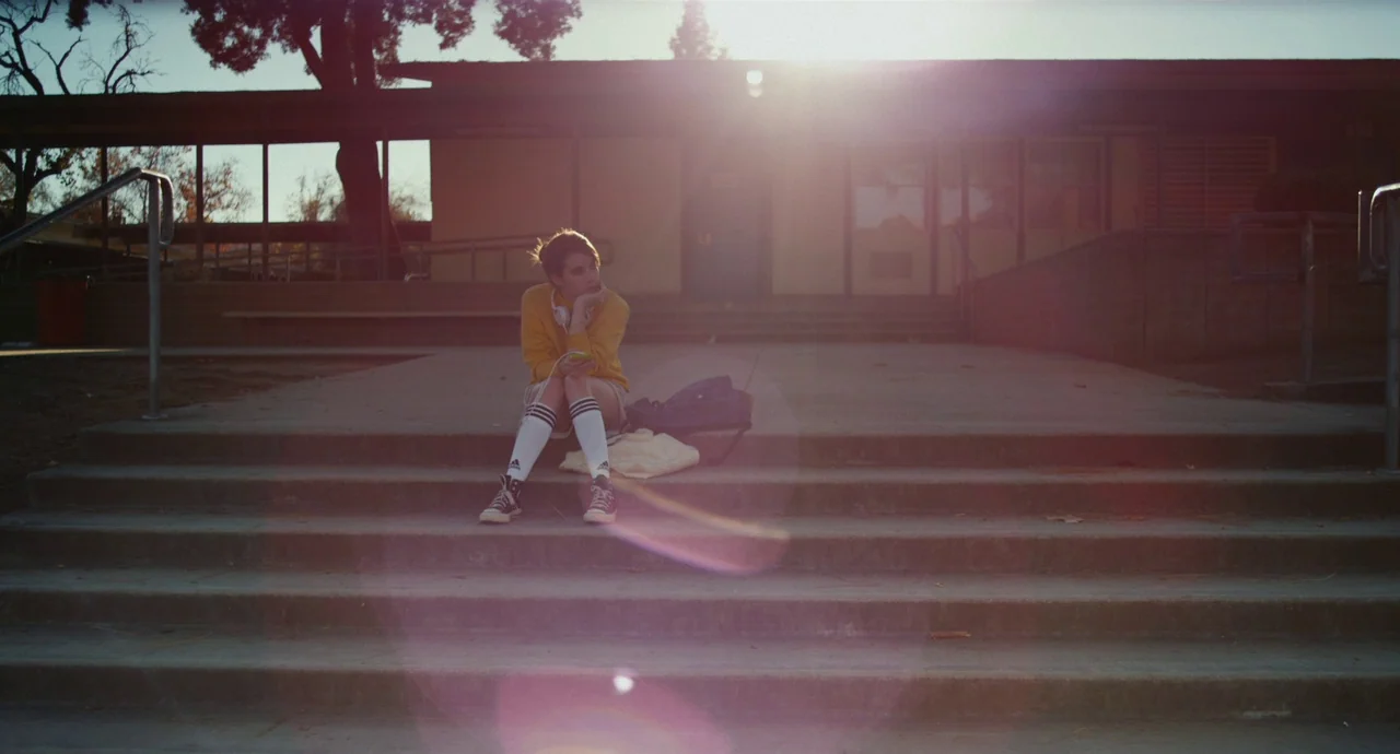Palo Alto (2013) movie still: wide — The concrete stairs and entrance of a school building during late afternoon. A young woman sits alone on the…
