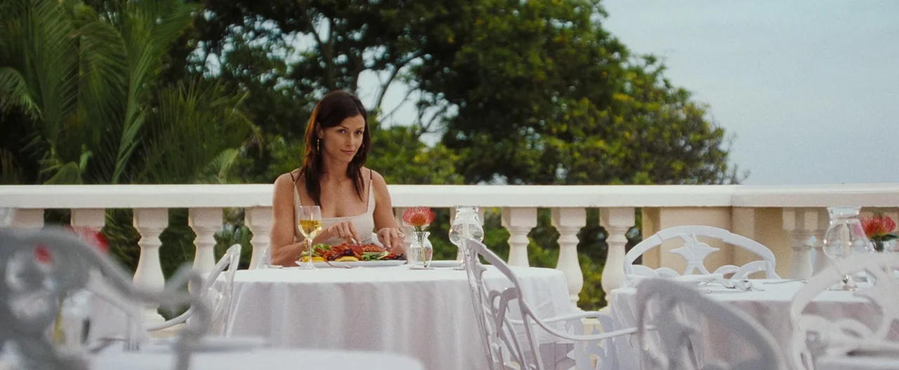 Lord of War (2005) movie still: medium — A woman sits at an outdoor table on a restaurant terrace with a balustrade. She is eating a meal while…