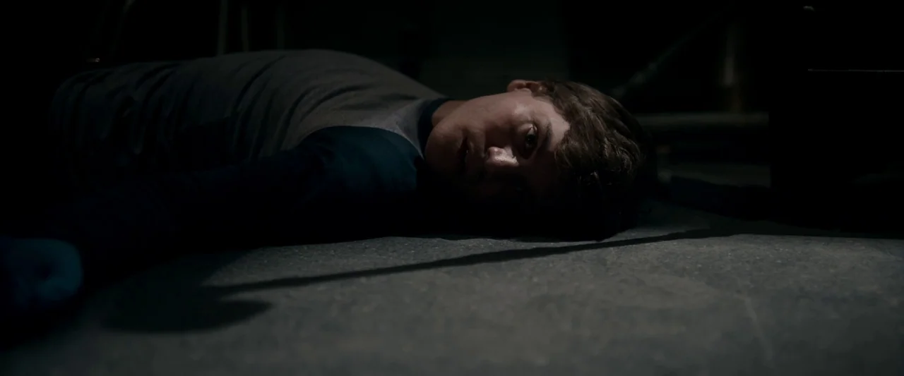 The Autopsy of Jane Doe (2016) movie still: closeup — A dark, dimly lit indoor space with a plain concrete floor. A young person lies prone on the…