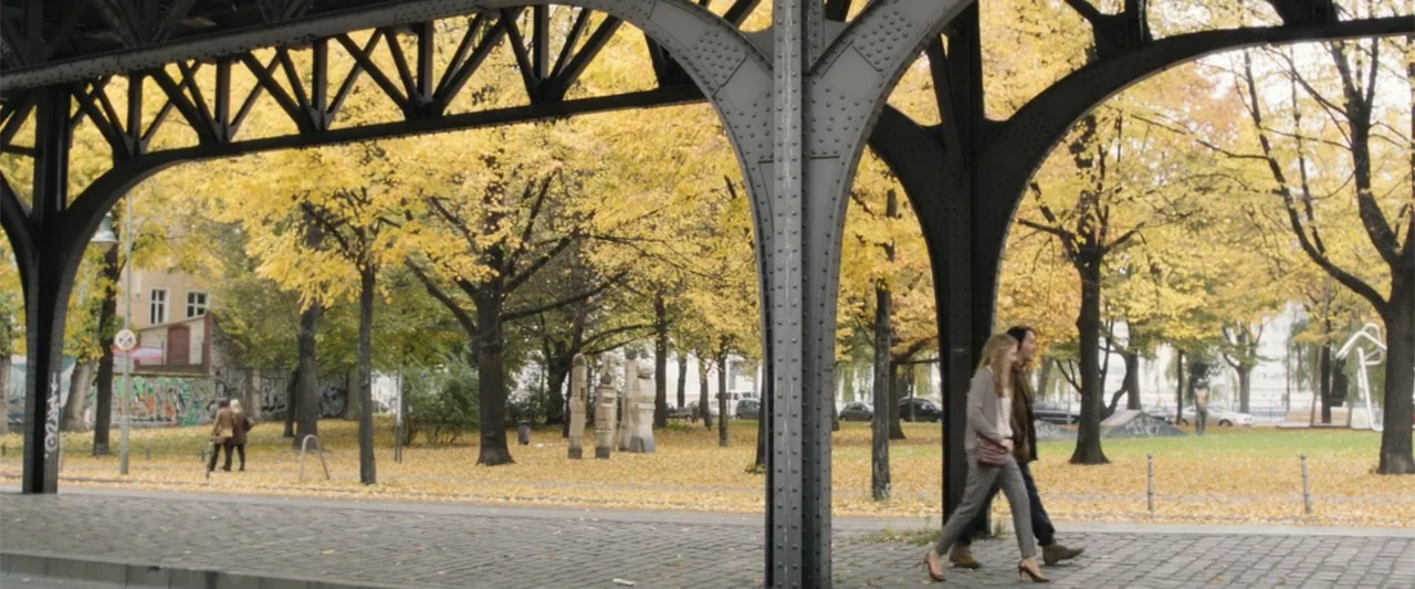Heartburn (1986) movie still: wide — An urban park in autumn features yellow trees and a large metal railway bridge. A man and a woman walk together…
