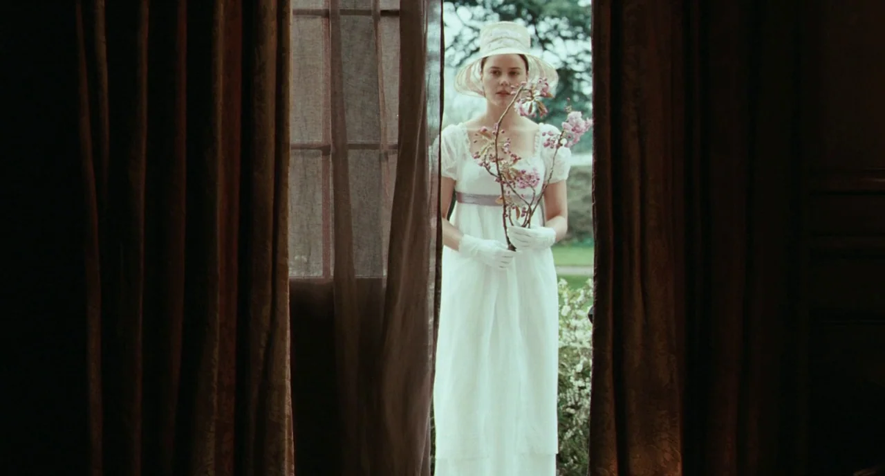 Bright Star (2009) movie still: medium — A woman stands in a doorway framed by dark brown heavy curtains. She holds a thin branch of pink cherry…